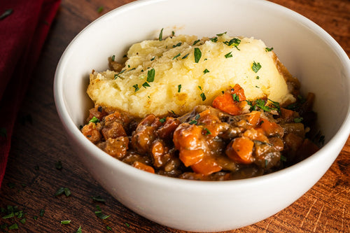 A white bowl filled with a hearty stew or chili-like dish topped with mashed potatoes and chopped parsley, set on a rustic wooden table with a wine glass in the background.