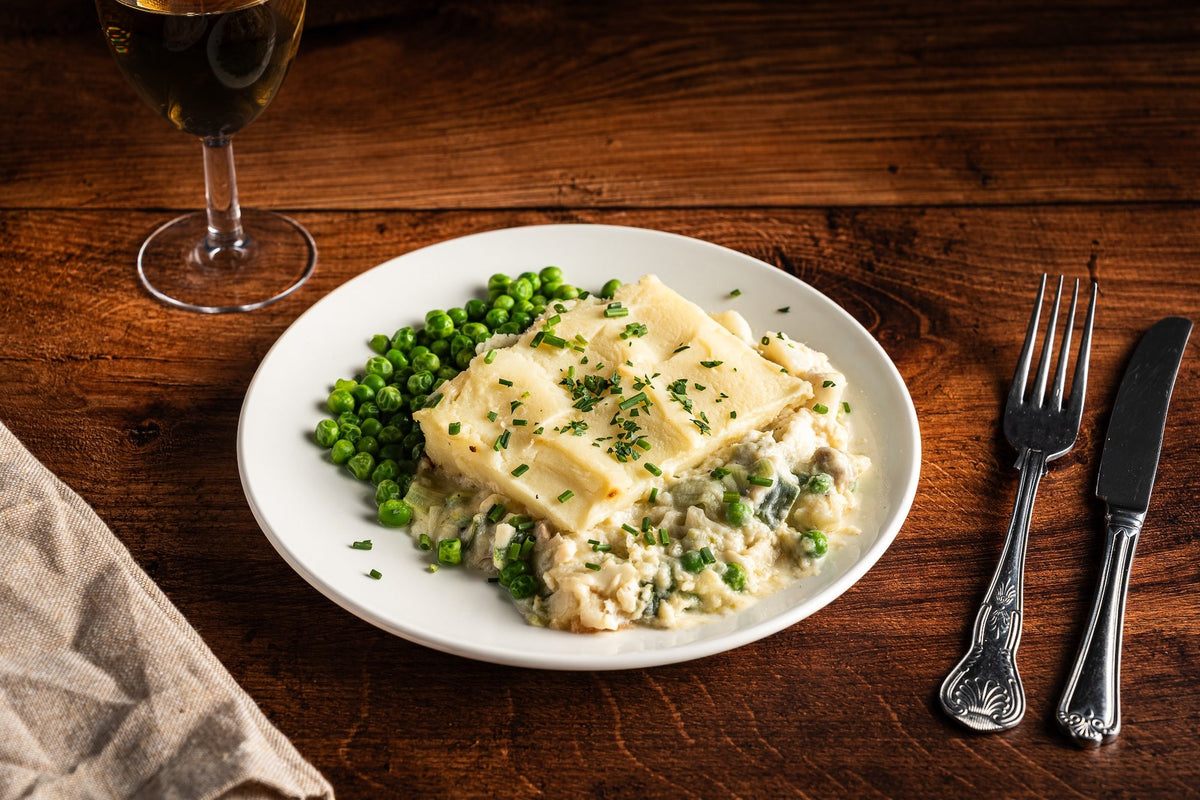 A plate of fish pie with a golden mashed potato topping and garden peas, on a dark wooden table with a glass of white wine and silver cutlery.