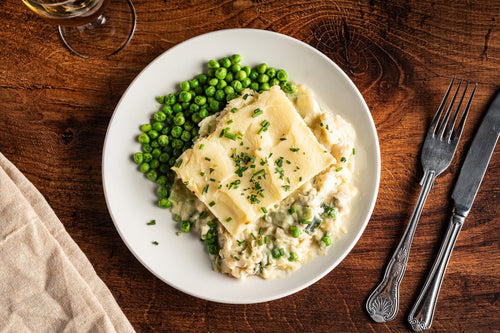 Overhead view of fish pie with a mashed potato topping scattered with chopped chives, creamy white fish and greens beneath, garden peas to one side.