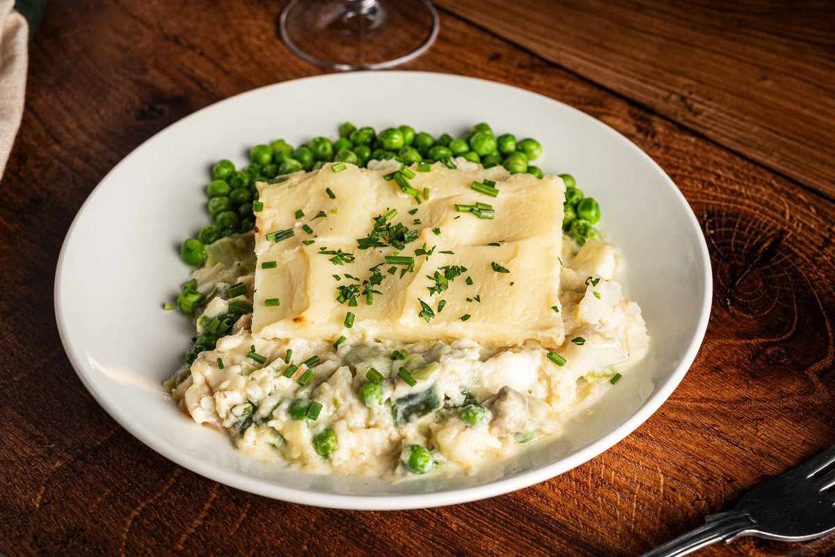 Fish pie with a mashed potato topping and garden peas on a white plate, with a linen napkin and a glass of white wine behind.