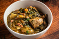 A white ceramic bowl filled with a hearty green vegetable and meat stew, set on a rustic dark wooden table with a dark green cloth in the background.