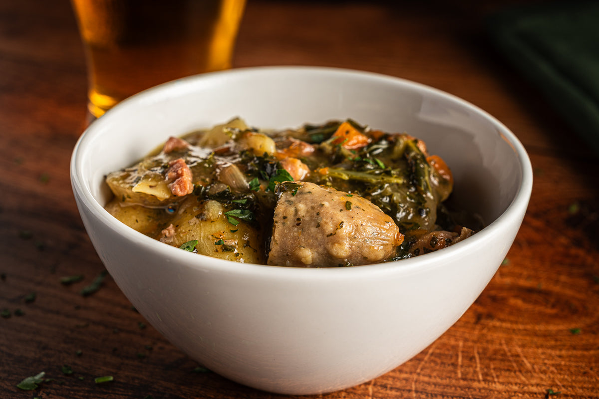 A close-up shot of a white ceramic bowl filled with a hearty stew or chowder containing potatoes, greens, and chunks of meat, set on a rustic wooden table with a blurred glass of beer in the background.