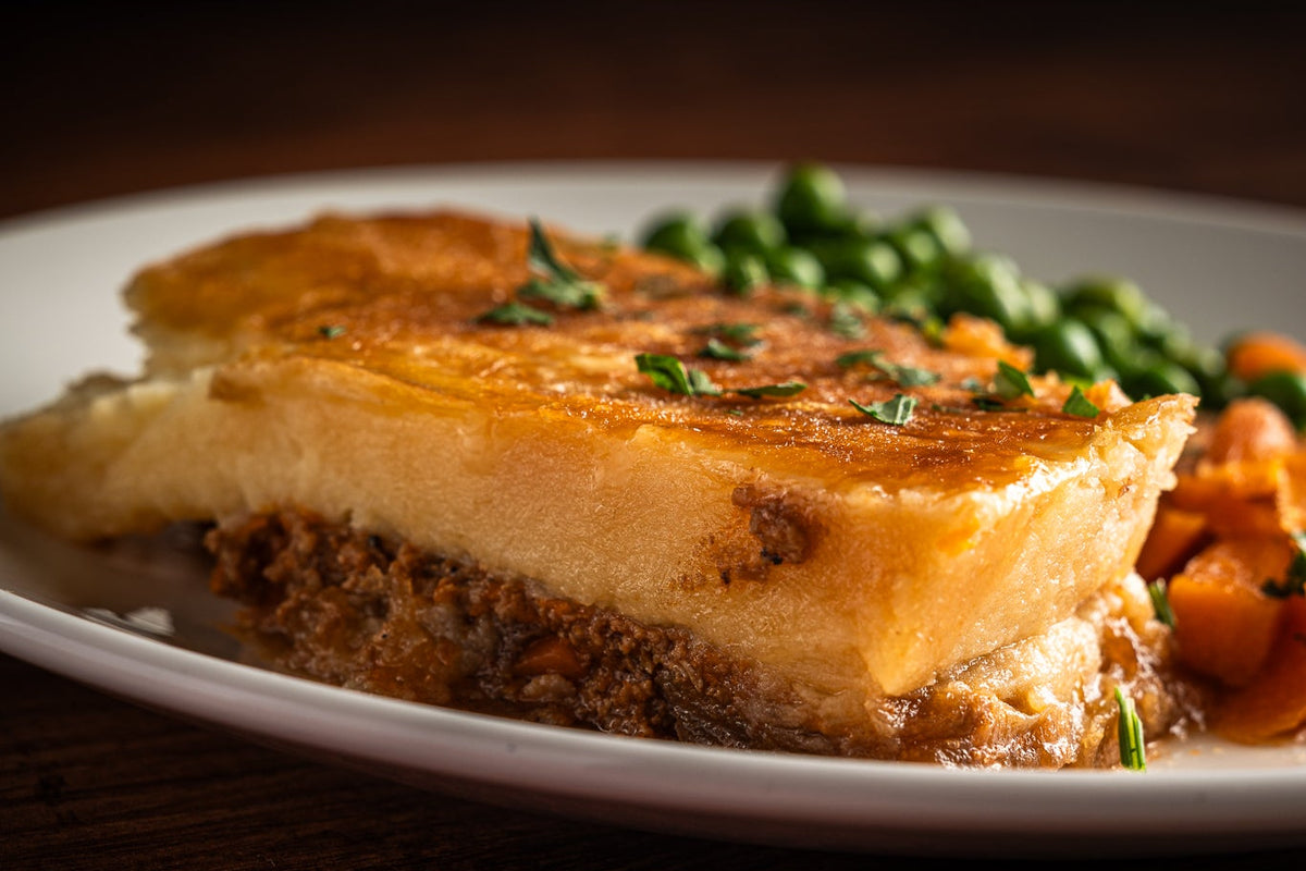 A close-up of cottage pie with a golden-brown top, served with green peas and carrots on a white plate against a dark wooden table.