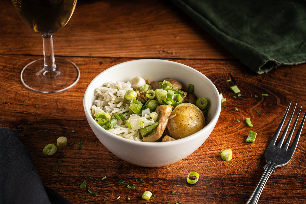 A white bowl of cooked rice with green onions, chicken, and a potato on a rustic wooden table, styled with a fork, a glass of white wine, and a dark green cloth.